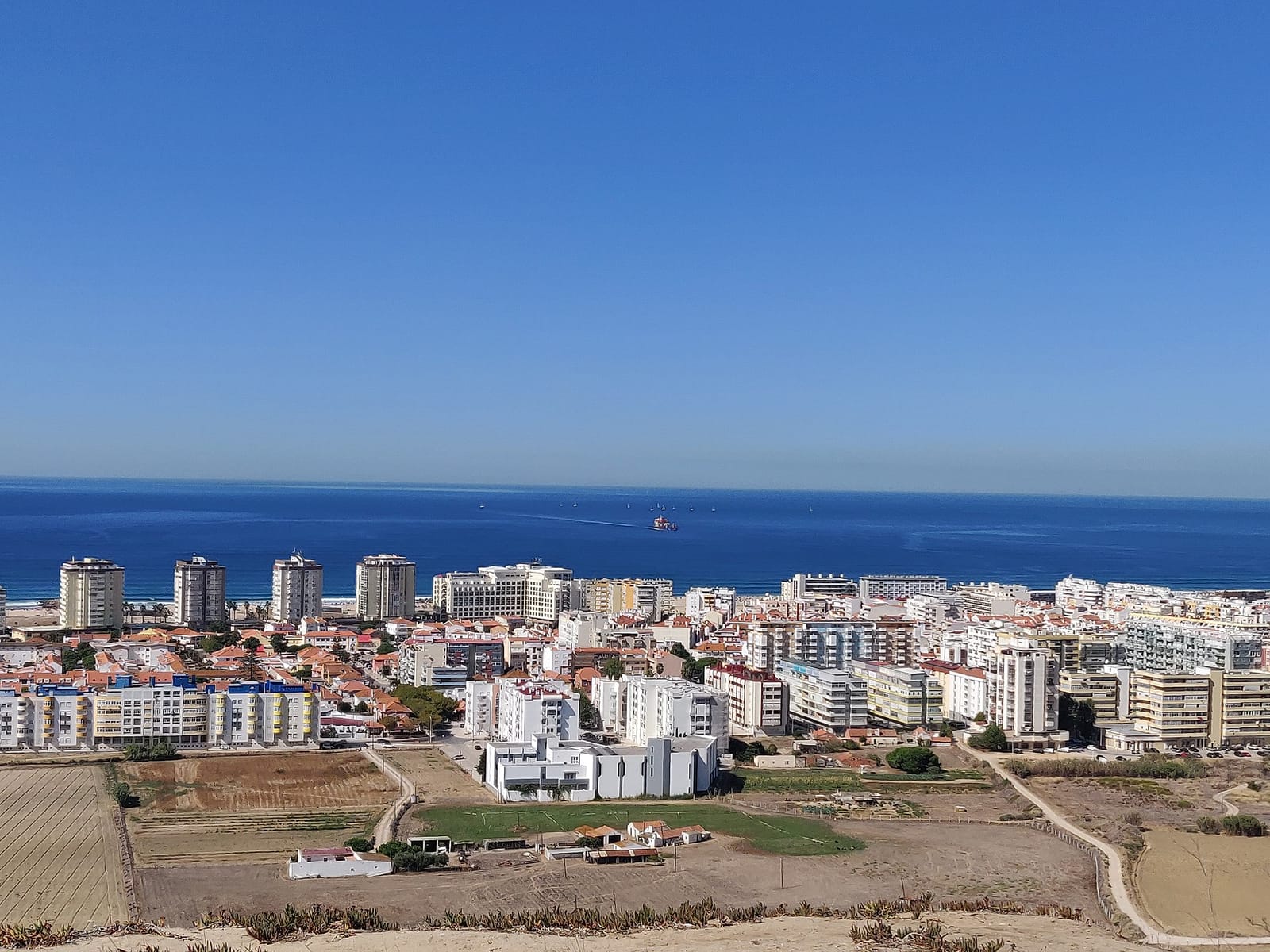 Vue aérienne de la Costa da Caparica : une ville dense de tours et d'immeubles blancs face à une longue plage atlantique, avec des champs maraîchers desséchés au premier plan.