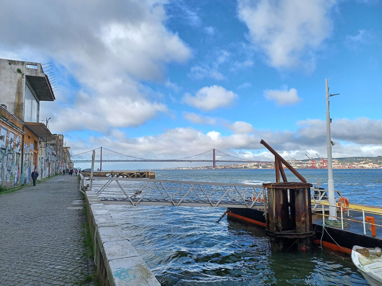 Quai pavé de Cacilhas bordé de bâtiments couverts de graffitis ; au loin, le pont du 25 Avril enjambe le Tage et la ville de Lisbonne s'étend sur l'autre rive.
