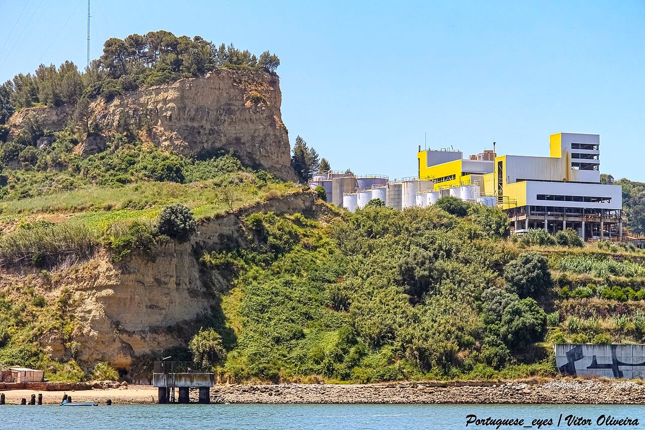 Falaise sèche et broussailleuse dominant le Tage à Almada, surmontée de silos et d'un complexe industriel jaune et blanc, sous un ciel sans nuage.