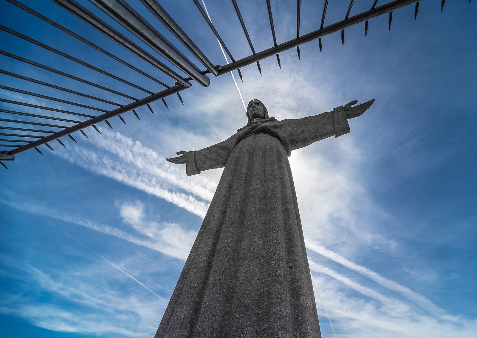 La statue du Cristo Rei d'Almada, vue en contre-plongée depuis sa base, bras écartés contre un ciel bleu strié de traînées d'avions.