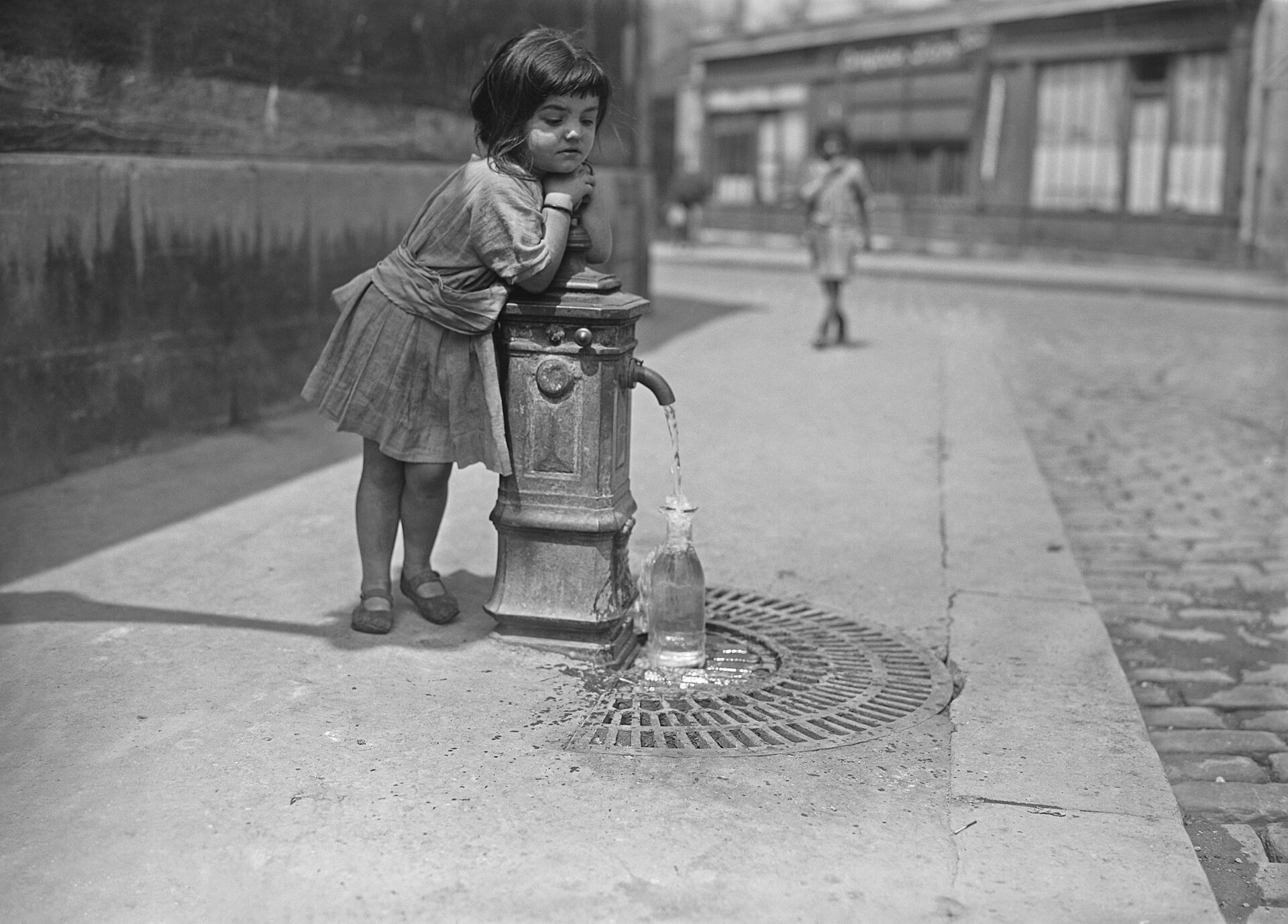 Photographie noir et blanc de 1921 : une fillette penchée sur une borne-fontaine de rue à Paris, un jour de forte chaleur